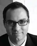 A black-and-white photograph of a man with dark hair and a cleft chin wearing glasses and looking at the viewer while smiling